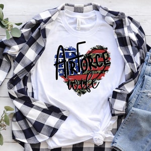 May include: White t-shirt with a red, white, and blue heart design that says "Air Force Wife". The heart is decorated with camouflage print and stars.