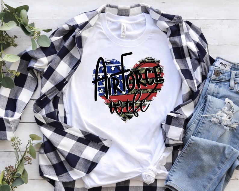 May include: White t-shirt with a red, white, and blue heart design that says "Air Force Wife". The heart is decorated with camouflage print and stars.