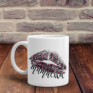 Pu&ograve; includere: Tazza in ceramica bianca con un design a labbra con stampa leopardata nera e rosa e la parola "mamacita" stampata sotto.
