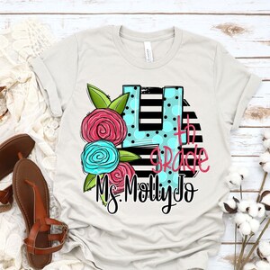 May include: A light gray t-shirt with a black and white striped background. The number 4 is in a blue circle with white polka dots. The text "4th Grade" is written in black. The text "Ms. Molly Jo" is written in black below the number 4. There are pink and blue flowers on the left side of the number 4.