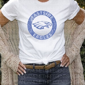 K&ouml;nnte beinhalten: Ein wei&szlig;es T-Shirt mit einem blauen, abgenutzten Kreis-Motiv, auf dem "Eastside Eagles" steht und ein blauer Adler im Kreis zu sehen ist.