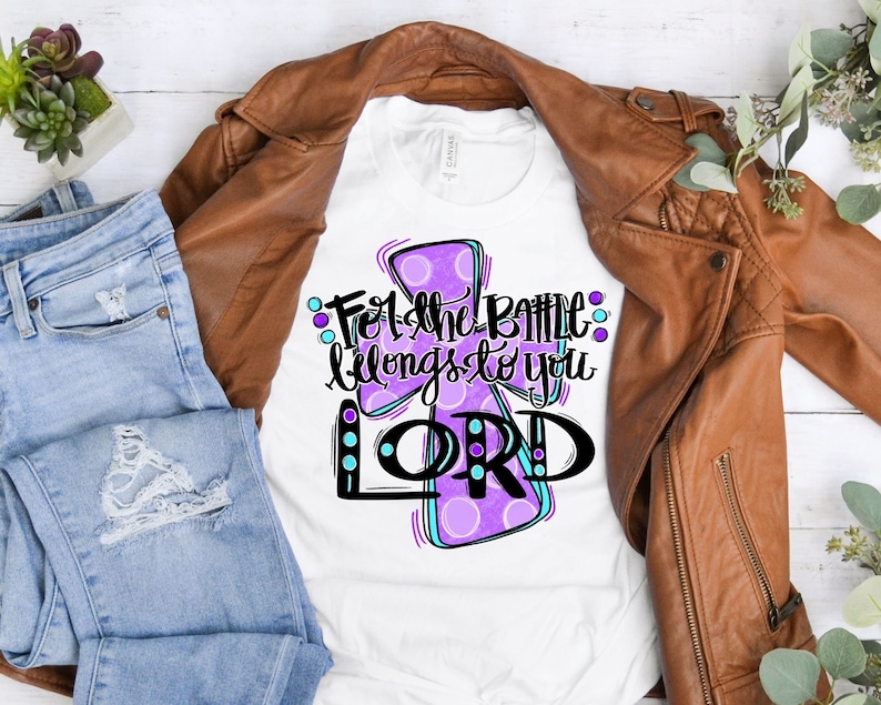 May include: White t-shirt with a purple and blue cross design. The text "For the battle belongs to you LORD" is written on the shirt.