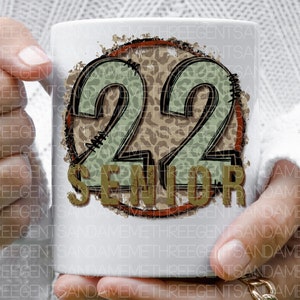 Puede incluir: Taza de cer&aacute;mica blanca con un dise&ntilde;o marr&oacute;n, rojo y verde. El dise&ntilde;o presenta el n&uacute;mero 22 con un fondo de estampado de leopardo y la palabra "Senior" en una fuente desgastada.