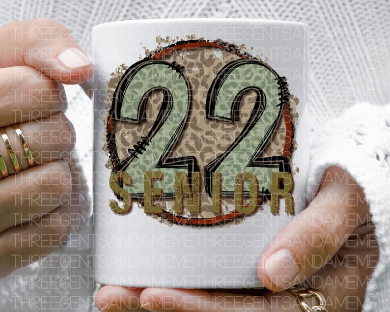 Puede incluir: Taza de cer&aacute;mica blanca con un dise&ntilde;o marr&oacute;n, rojo y verde. El dise&ntilde;o presenta el n&uacute;mero 22 con un fondo de estampado de leopardo y la palabra "Senior" en una fuente desgastada.
