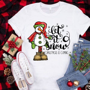 May include: White t-shirt with a cartoon snowman wearing a red scarf and green mittens. The text "Let it snow" is written in black cursive with snowflakes. The text "Christmas is coming" is written in black below the snowman.