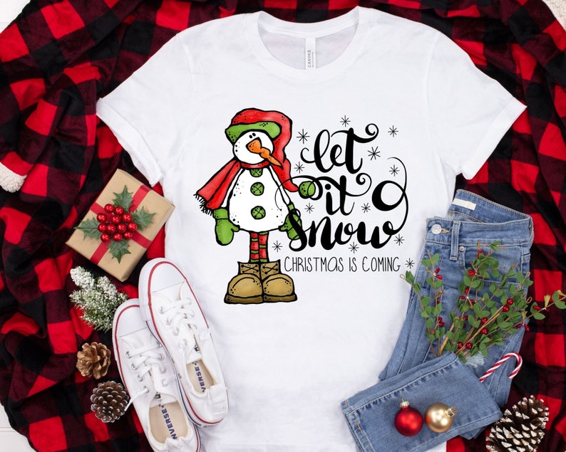 May include: White t-shirt with a cartoon snowman wearing a red scarf and green mittens. The text "Let it snow" is written in black cursive with snowflakes. The text "Christmas is coming" is written in black below the snowman.