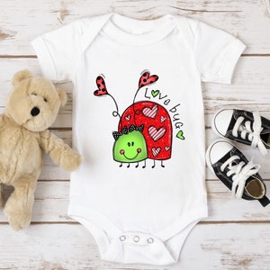 K&ouml;nnte beinhalten: Wei&szlig;er Baby-Strampler mit einem roten und gr&uuml;nen Marienk&auml;fer-Design. Der Marienk&auml;fer hat Herzen auf dem R&uuml;cken und der Text "Love bug" steht daneben.