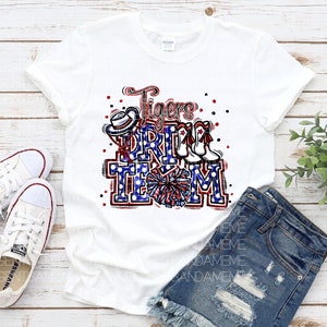 May include: White t-shirt featuring a red, white and blue graphic that reads "Tigers Drill Team". The graphic includes a cheerleader's pom pom, a hat and boots.