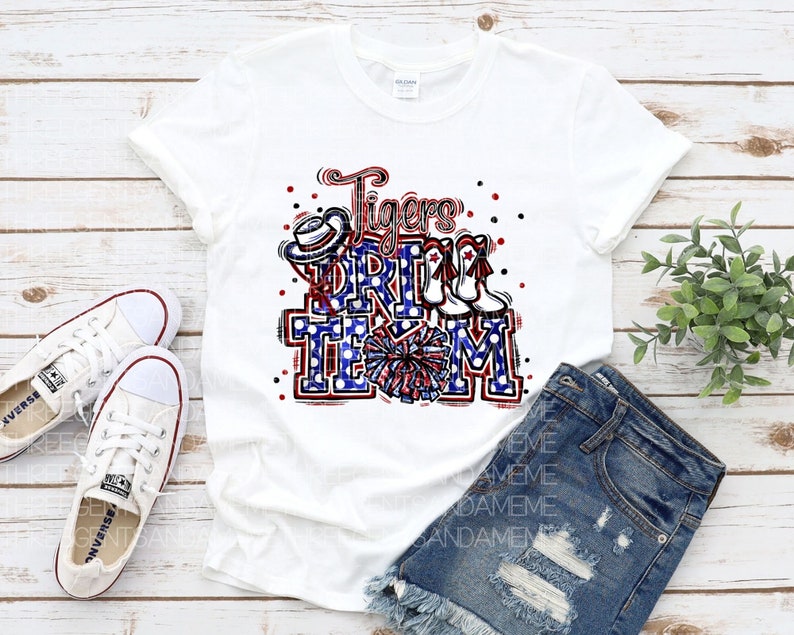 May include: White t-shirt featuring a red, white and blue graphic that reads "Tigers Drill Team". The graphic includes a cheerleader's pom pom, a hat and boots.