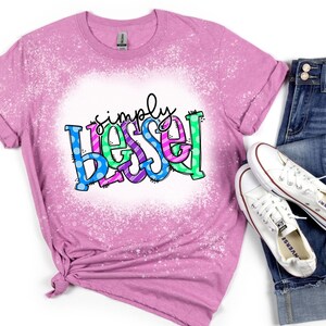 May include: A pink t-shirt with a bleach-splattered design featuring the text "simply blessed" in colorful letters with polka dots.