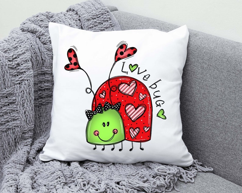 K&ouml;nnte beinhalten: Ein wei&szlig;es Kissen mit einem roten und gr&uuml;nen Marienk&auml;fer-Design. Der Marienk&auml;fer hat schwarze Augen und eine schwarze Schleife. Der Text "Love Bug" ist in gr&uuml;nen Buchstaben auf dem Kissen geschrieben.