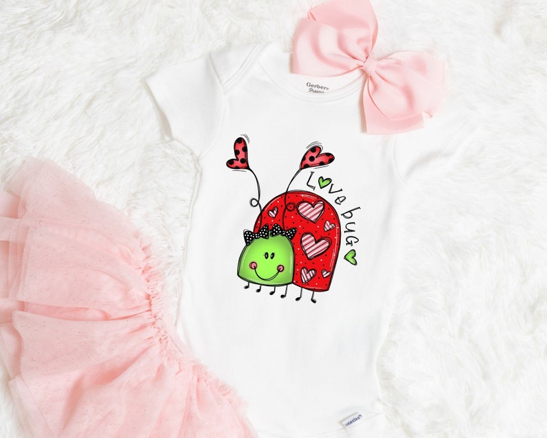 K&ouml;nnte beinhalten: Wei&szlig;er Strampler mit einem roten und gr&uuml;nen Marienk&auml;fer-Design. Der Marienk&auml;fer hat Herzen auf dem R&uuml;cken und der Text "Love Bug" steht an der Seite.