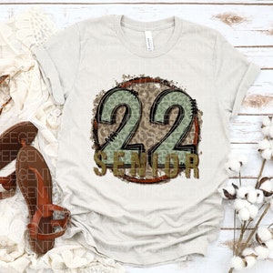 Puede incluir: Una camiseta blanca con un dise&ntilde;o circular marr&oacute;n y verde que presenta el n&uacute;mero 22 y la palabra "Senior".