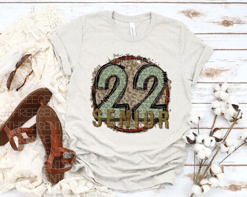 Puede incluir: Una camiseta blanca con un dise&ntilde;o circular marr&oacute;n y verde que presenta el n&uacute;mero 22 y la palabra "Senior".