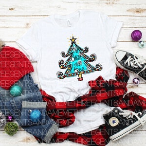 May include: A white t-shirt featuring a colourful Christmas tree design and the text "Oh Christ was a tree". The shirt is surrounded by a red and black checked shirt, blue jeans, a red knitted hat, and some Christmas decorations.