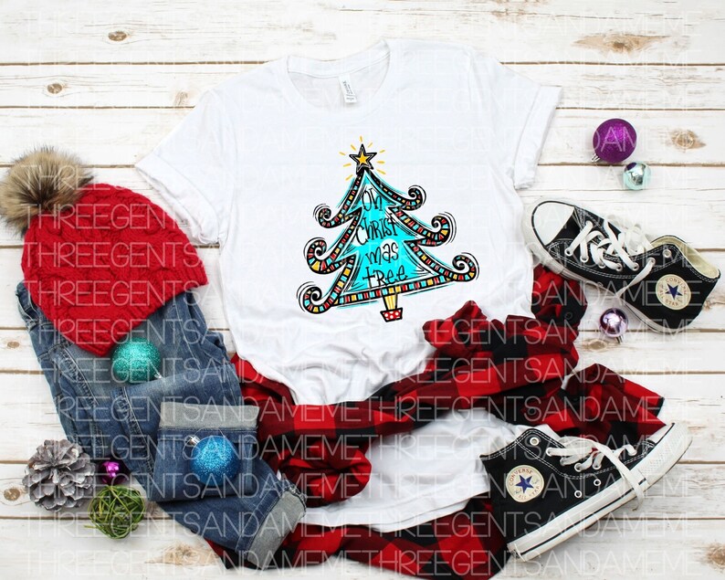 May include: A white t-shirt featuring a colourful Christmas tree design and the text "Oh Christ was a tree". The shirt is surrounded by a red and black checked shirt, blue jeans, a red knitted hat, and some Christmas decorations.