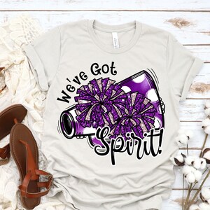 Op de afbeelding: Een wit T-shirt met een paarse en witte cheerleading pompon-afbeelding en de tekst "We've Got Spirit!" erop gedrukt.