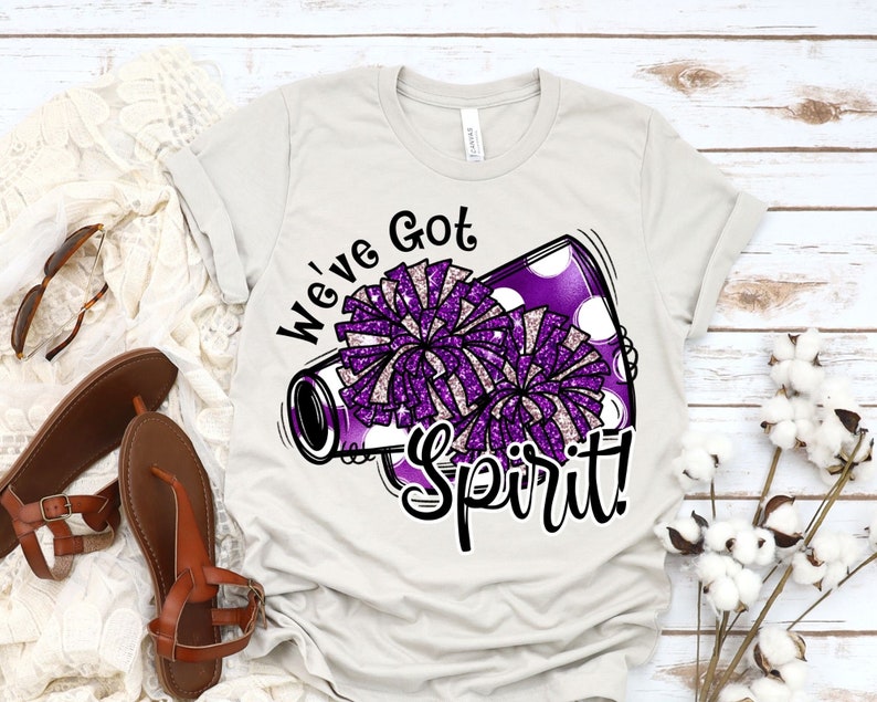 Op de afbeelding: Een wit T-shirt met een paarse en witte cheerleading pompon-afbeelding en de tekst "We've Got Spirit!" erop gedrukt.