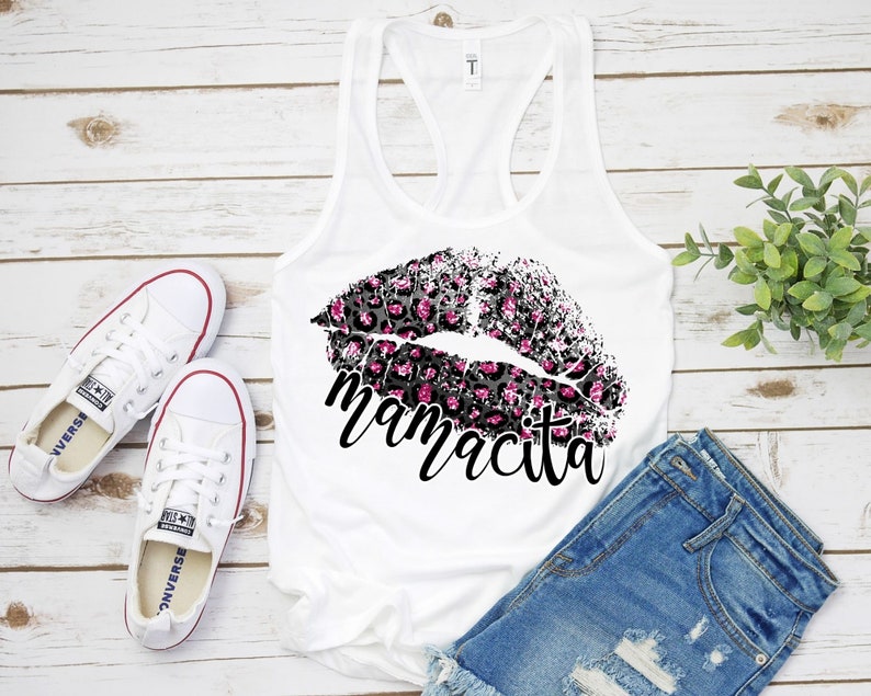 Pu&ograve; includere: Canotta bianca con un designo di labbra a stampa leopardata nera e rosa e la parola "mamacita" stampata sotto. La canotta &egrave; appoggiata su una superficie di legno bianco con un paio di scarpe da ginnastica bianche e pantaloncini di jeans blu.