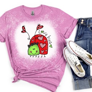 K&ouml;nnte beinhalten: Ein pinkfarbenes T-Shirt mit einem Bleiche-Spritzer-Design, das ein Cartoon-K&auml;ferpaar mit Herzen und dem Text "Love bug" zeigt.