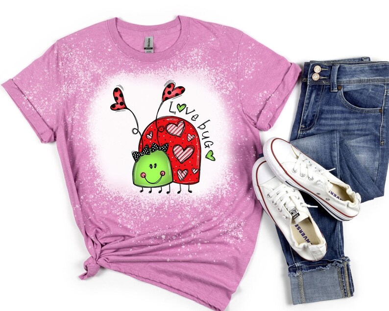 K&ouml;nnte beinhalten: Ein pinkfarbenes T-Shirt mit einem Bleiche-Spritzer-Design, das ein Cartoon-K&auml;ferpaar mit Herzen und dem Text "Love bug" zeigt.