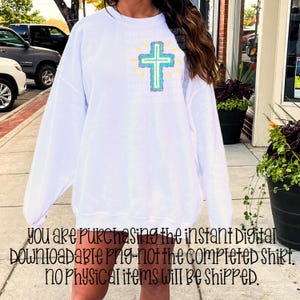 May include: White long-sleeve sweatshirt featuring a colorful cross design on the front. The cross is outlined in turquoise and teal, with yellow rays emanating from behind. The text on the sweatshirt is partially visible.