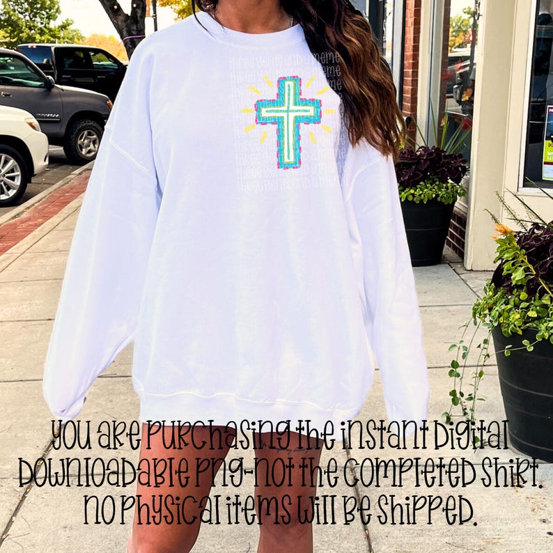 May include: White long-sleeve sweatshirt featuring a colorful cross design on the front. The cross is outlined in turquoise and teal, with yellow rays emanating from behind. The text on the sweatshirt is partially visible.