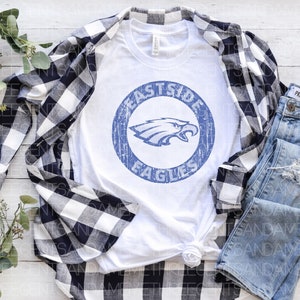 K&ouml;nnte beinhalten: Wei&szlig;es T-Shirt mit einem blauen und wei&szlig;en, abgenutzten Kreis-Motiv, auf dem "Eastside Eagles" steht. Das Shirt liegt auf einer wei&szlig;en Holzoberfl&auml;che mit einem schwarz-wei&szlig; karierten Hemd und einer blauen Jeans.