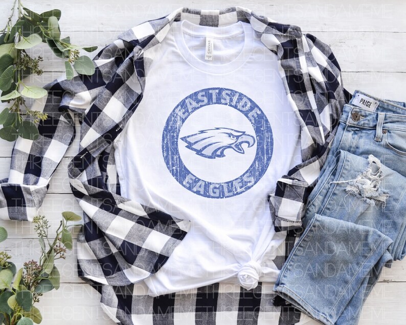 K&ouml;nnte beinhalten: Wei&szlig;es T-Shirt mit einem blauen und wei&szlig;en, abgenutzten Kreis-Motiv, auf dem "Eastside Eagles" steht. Das Shirt liegt auf einer wei&szlig;en Holzoberfl&auml;che mit einem schwarz-wei&szlig; karierten Hemd und einer blauen Jeans.