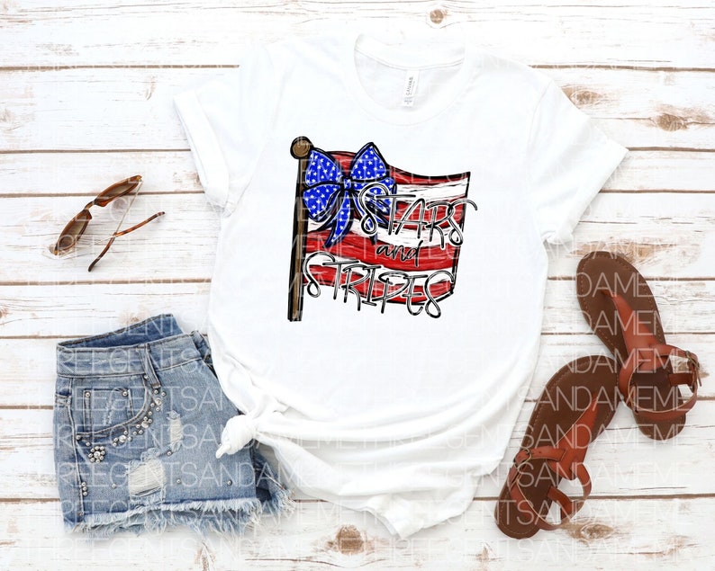 May include: A white t-shirt with a red, white, and blue American flag design. The flag has a blue bow on top and the text "Stars and Stripes" written on it.