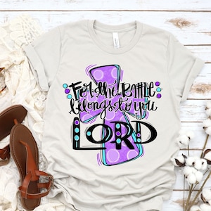 May include: A light gray t-shirt with a purple and blue cross design. The cross has white polka dots and the text "For the battle belongs to you LORD" is written in black.