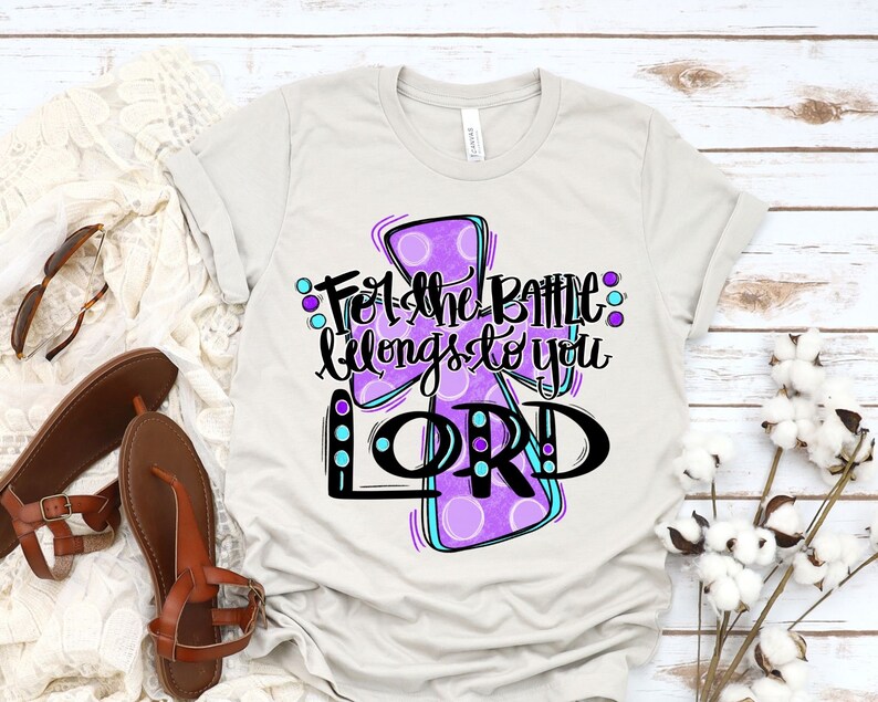 May include: A light gray t-shirt with a purple and blue cross design. The cross has white polka dots and the text "For the battle belongs to you LORD" is written in black.