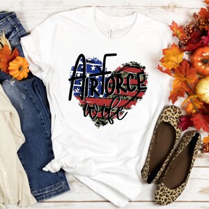 May include: White t-shirt with a red, white, and blue heart design that says "Air Force Wife". The heart is distressed and has a camouflage pattern.