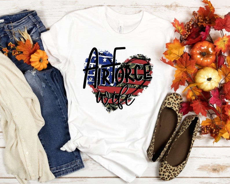 May include: White t-shirt with a red, white, and blue heart design that says "Air Force Wife". The heart is distressed and has a camouflage pattern.