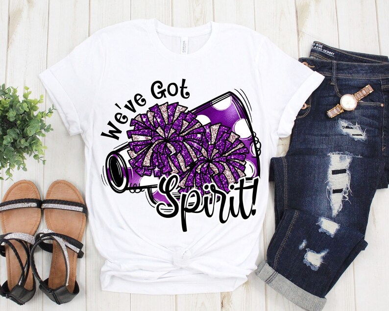 Op de afbeelding: Wit T-shirt met een afbeelding van een paarse en witte glitterpompon en een megafoon. De tekst "We've Got Spirit!" is op het shirt gedrukt.