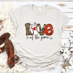May include: A white t-shirt with a graphic design of the word "LOVE" with a baseball in the center. The letters are decorated with leopard print, stripes, and red glitter. The text "for the love of the game" is written below the graphic.