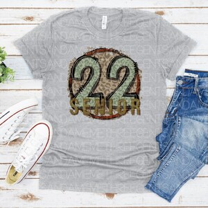 Puede incluir: Camiseta gris con un gr&aacute;fico circular desgastado que presenta el n&uacute;mero 22 en dorado con un estampado de leopardo verde y marr&oacute;n. El texto "Senior" est&aacute; escrito en dorado debajo del n&uacute;mero.