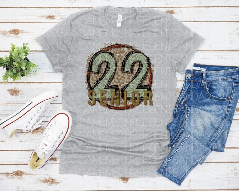 Puede incluir: Camiseta gris con un gr&aacute;fico circular desgastado que presenta el n&uacute;mero 22 en dorado con un estampado de leopardo verde y marr&oacute;n. El texto "Senior" est&aacute; escrito en dorado debajo del n&uacute;mero.
