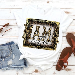 May include: White t-shirt with a gold and black distressed frame featuring a leopard print design and the text "Mamacita".