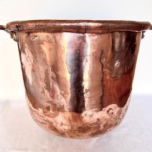 May include: A large, round, hammered copper pot with a wide, rolled rim and two small metal side handles. The exterior surface features a polished, reflective finish with visible signs of wear, including minor scratches and surface variations.