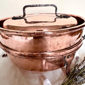 May include: A copper cooking pot with a lid and a handle. The pot has a rounded shape and sits on three small feet. The handle is made of a darker metal. A bundle of dried lavender is in the foreground.