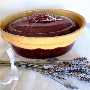 May include: A vintage ceramic casserole dish with a dark brown lid and a tan and brown base. The dish is oval-shaped with handles. A bundle of dried lavender tied with a white ribbon is next to the dish.