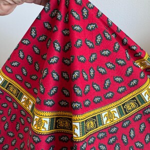 May include: A red fabric with a paisley pattern and a decorative border. The paisley shapes are black, white, and yellow on a red background. The border features yellow, green, and white stripes with ornate designs.