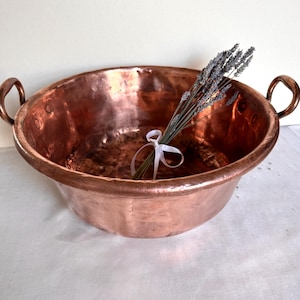 May include: A large, antique copper bowl with two loop handles. The bowl has a wide, shallow design and a rich, reddish-brown color. A bundle of dried lavender tied with a white ribbon rests inside the bowl, adding a touch of rustic charm.