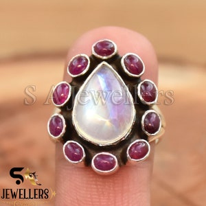 May include: A silver ring with a pear-shaped moonstone surrounded by a halo of small red gemstones.