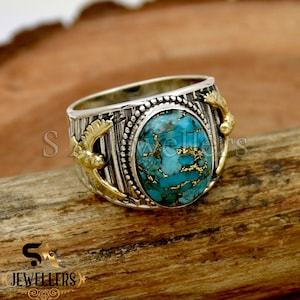 May include: A silver ring with a turquoise stone and gold accents. The ring has a detailed design with two wings on either side of the stone.