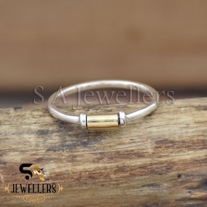 May include: A silver ring with a gold-coloured central bar. The ring has a simple, elegant design, suitable for everyday wear. The ring is displayed on a wooden surface. The text "S Jewellers" is visible.
