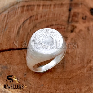 May include: A silver signet ring with a heart design. The heart is surrounded by a crown and rays of light. The ring is on a wooden surface.
