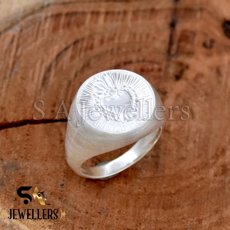 May include: A silver signet ring with a heart design. The heart is surrounded by a crown and rays of light. The ring is on a wooden surface.
