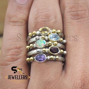 May include: A stack of silver rings with gold accents and colorful gemstones. The rings feature oval-shaped stones in yellow, blue, green, red, and purple. The rings are on a person's finger.